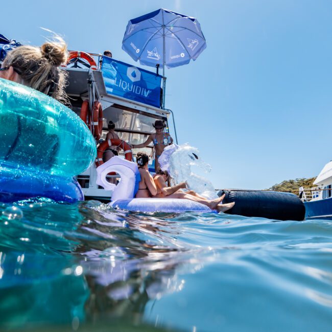 People lounging on inflatable floats and a boat in a calm body of water. The boat has a large blue umbrella, a visible logo, and is creating a social atmosphere. The sun is shining brightly, with a clear blue sky in the background.