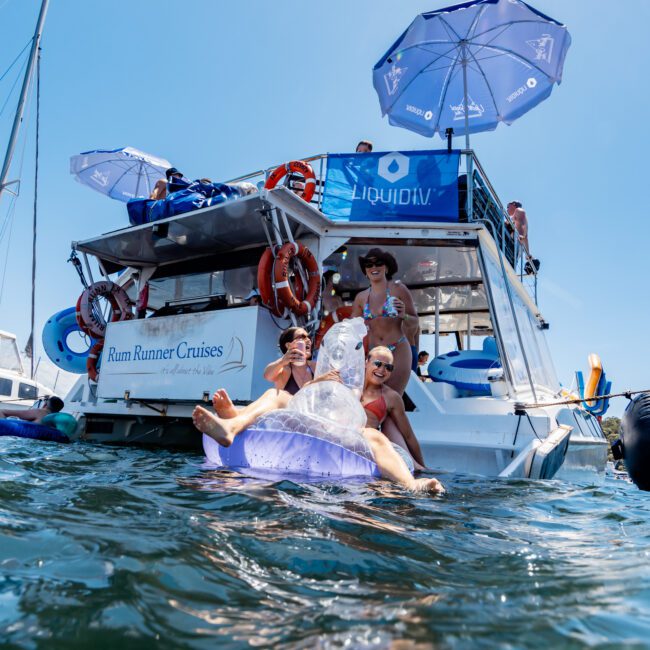 Group of people enjoying a sunny day on a boat named "Rum Runner Cruises," with some lounging under umbrellas and others sitting on inflatable floaties in the water. The scene is lively and festive, with a clear blue sky above.