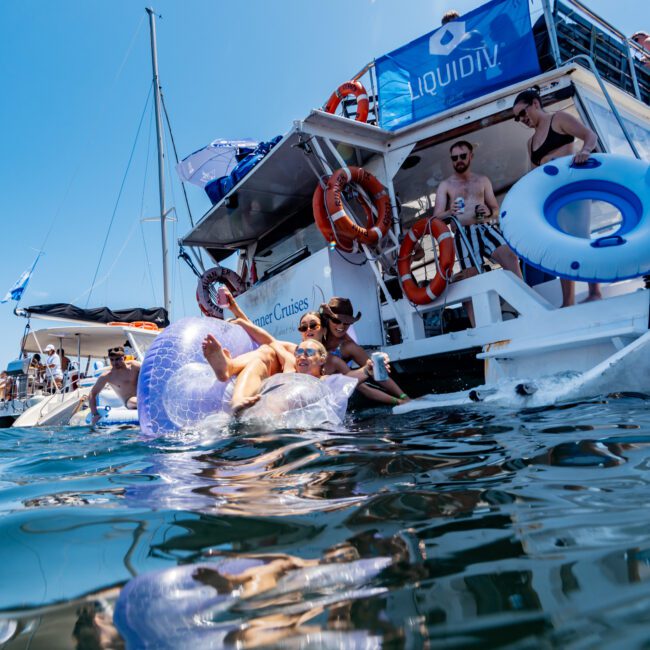 People are relaxing and enjoying on a yacht and inflatable tubes in blue water. Some are under an umbrella, others on a tube in the water. The scene is lively under a sunny sky.