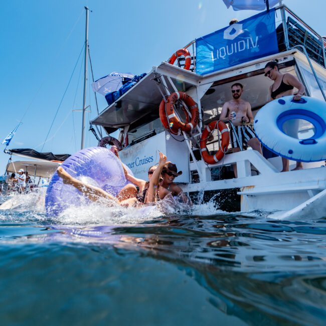 A group of people enjoy a sunny day on a boat, with some swimming in the water using inflatable rings. The boat is equipped with life buoys and shaded areas under blue umbrellas. The atmosphere is lively and cheerful.