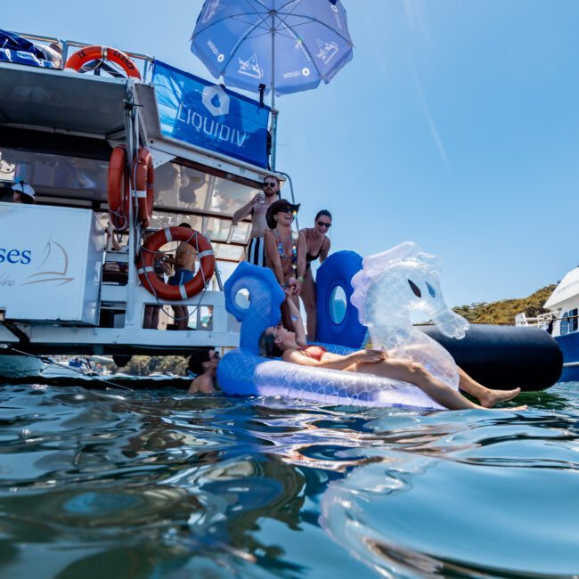 People relaxing on a yacht and a giant inflatable in the water. An umbrella shades part of the boat. The sky is clear and the shoreline is visible in the background.