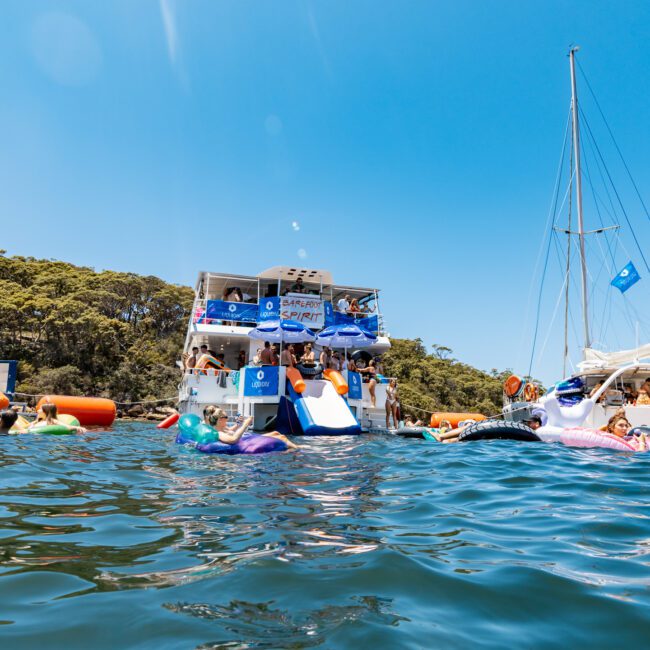 A group of people enjoys a sunny day on the water near several anchored boats. Some are on large inflatable loungers, while others play in the water. A nearby forested shoreline and a clear blue sky create a serene backdrop.