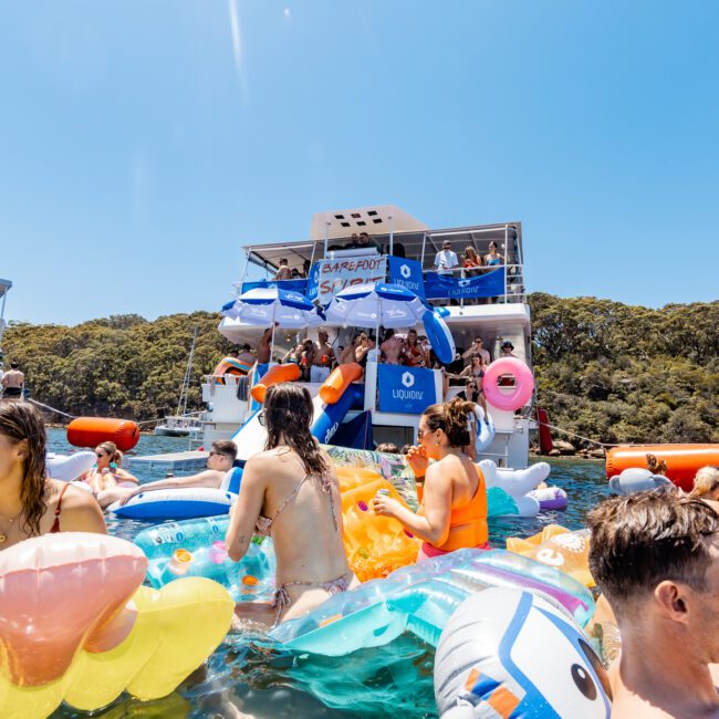 People enjoying a sunny day in the water with colorful inflatables near a blue and white boat. The background features a forested shoreline. Some individuals are on the boat, which has a slide leading into the water.