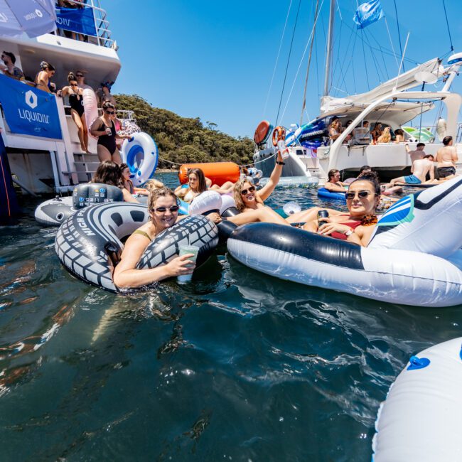 People are enjoying a sunny day on the water, sitting on inflatable rafts near boats. They are wearing swimsuits and sunglasses. The scene is lively, with clear skies and trees visible in the background.