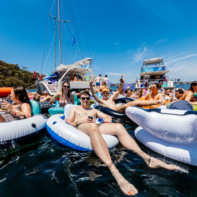 People relaxing on inflatable floats in the water, surrounded by boats. It's a sunny day, and the group appears to be enjoying a social event with drinks. Lush greenery and clear blue skies are in the background.