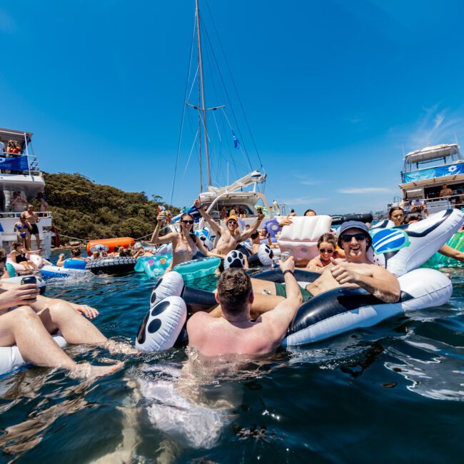 A lively scene on the water with people on inflatable rafts enjoying a sunny day. Boats are docked nearby, and participants are interacting, laughing, and holding drinks. The sky is clear, and trees are visible in the background.