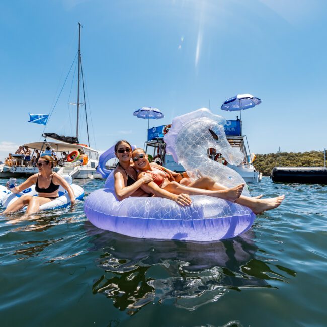 People relaxing on inflatable pool floats in the water near a yacht. They are enjoying a sunny day, with the blue sky and trees in the background. There are umbrellas on the yacht and various inflatable floaties around them.