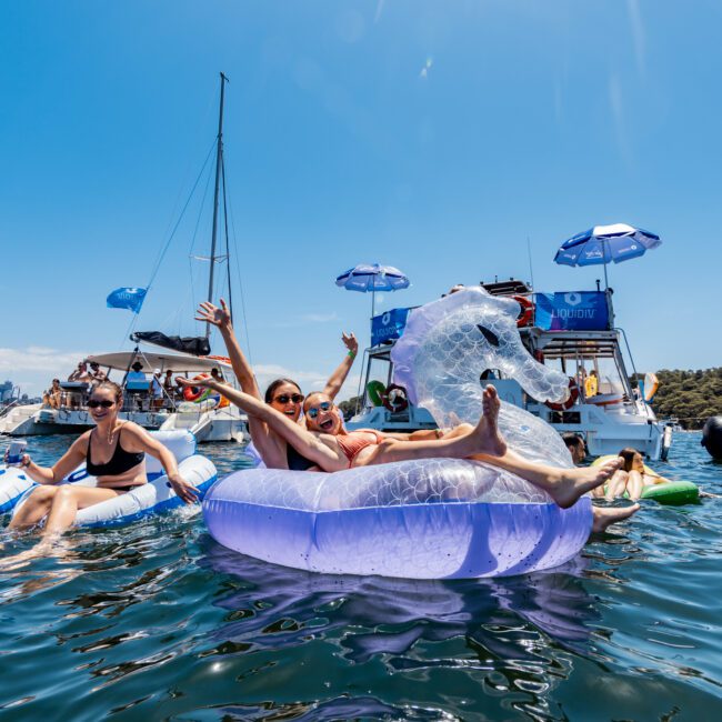 People relax on inflatable floats in the water, enjoying a sunny day. Boats are anchored nearby with umbrellas providing shade. The atmosphere is festive and summery.