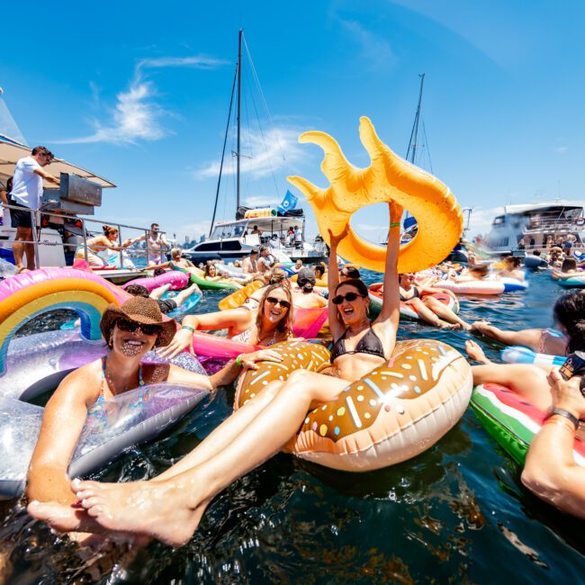 People relaxing on colorful inflatables in the water, surrounded by yachts. They're enjoying a sunny day, with one holding a large inflatable shaped like a sun. The scene is lively and festive, with clear blue skies overhead.