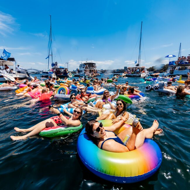 A lively group of people relax on colorful floaties in a sunny, crowded bay, surrounded by anchored boats. The scene is festive, with individuals enjoying the water and warm weather in swimsuits and sunglasses.