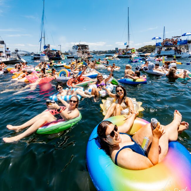 A lively scene of people floating on colorful inflatables in a sunny, crowded bay with boats in the background. The group is enjoying drinks and socializing, while the clear blue sky stretches overhead.