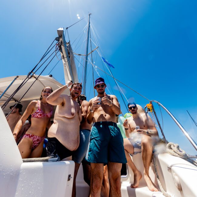 A group of people smiles and poses on a sailboat under a clear blue sky. They are wearing swimwear and sunglasses. The sun shines brightly overhead, creating a vibrant and lively atmosphere on the boat.