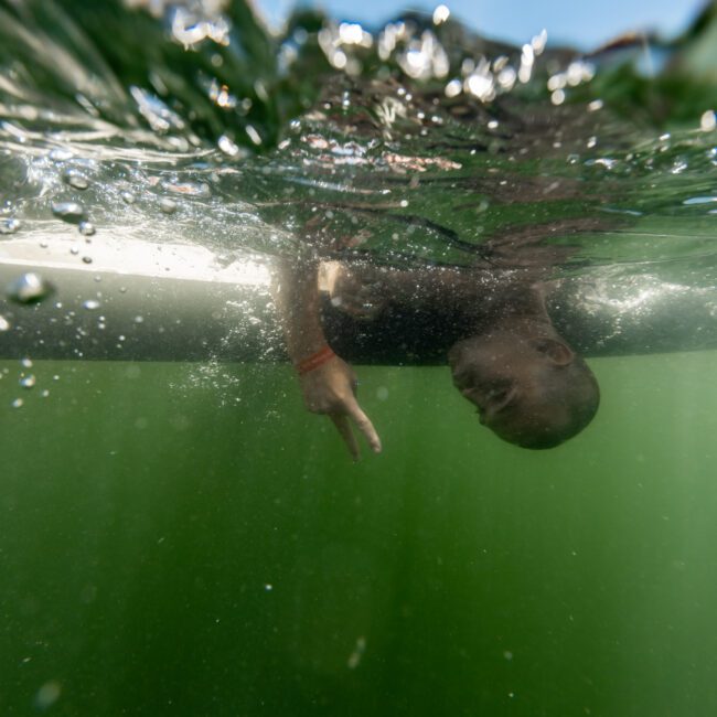 A person wearing a black shirt is floating upside down in green water, partially submerged. Their face and hand are visible beneath the water's surface. The water reflects light from above.