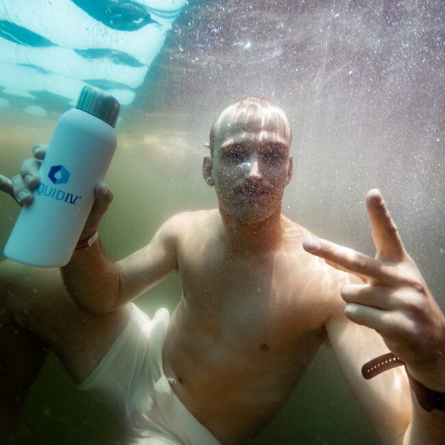 A man underwater wearing white swim trunks holds up a white water bottle with a blue logo. He gestures with two fingers on his other hand. Bubbles surround him in a murky greenish environment. A small logo reads "The Yacht Social Club.