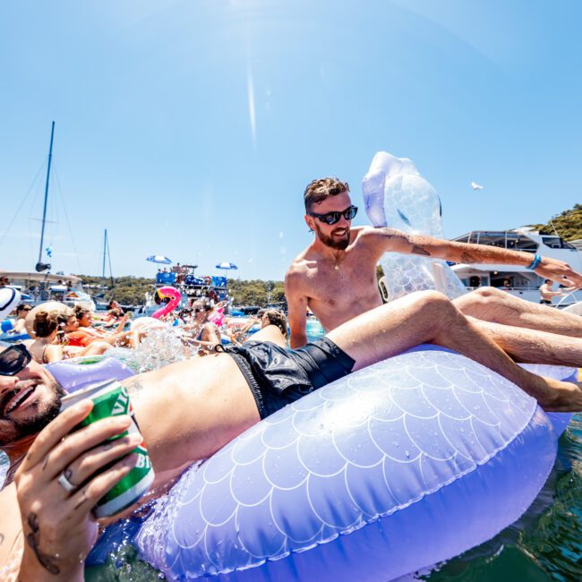 A group of people enjoying a sunny day in the water. Two men in sunglasses recline on inflatable floats, with one holding a drink. Behind them, a crowd mingles among various floats. Yachts and a clear sky are visible in the background.