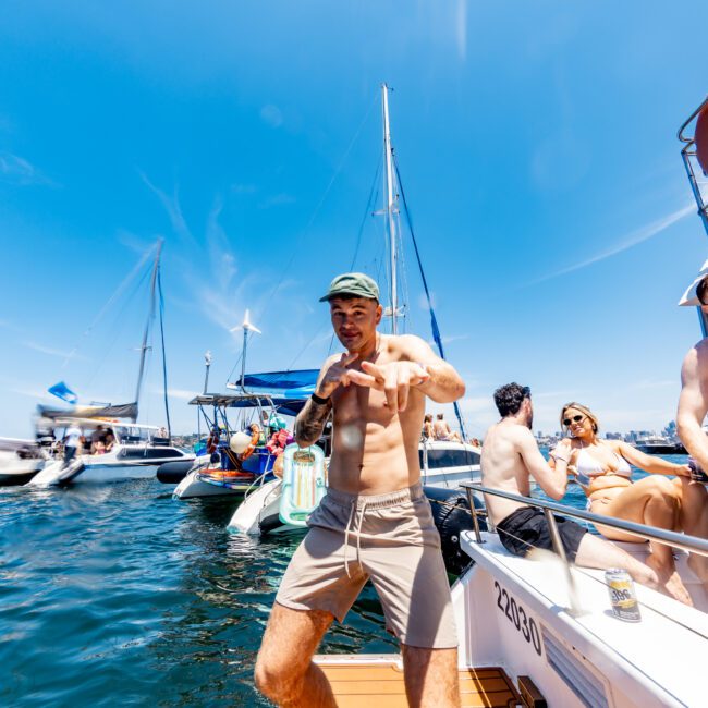 A shirtless man in shorts poses energetically on a boat deck surrounded by other boats and people under a clear blue sky. Others are relaxing on the boat, chatting, and sunbathing.