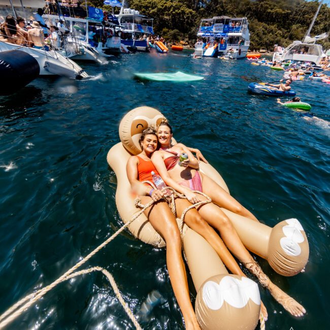 Two people are lying on a large inflatable shaped like a lounging figure, floating in a sunny marina filled with boats and other floaties. They smile and relax, holding drinks, while crowds enjoy the water around them.