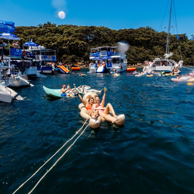 People are relaxing on inflatable floats linked together in a vibrant blue sea. Boats are anchored nearby, with more people gathering and socializing on them. Lush green trees line the shore under a clear sky.