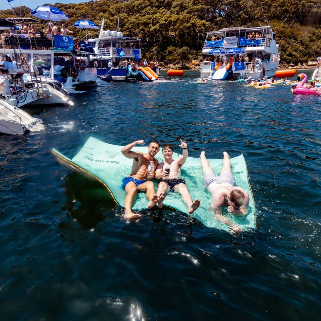 Three people relax on a floating mat in a lively water scene with boats and others enjoying the sun. Trees line the distant shore, and the water reflects the bright, sunny day. The atmosphere is festive and social.