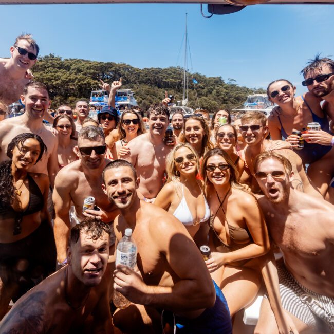 A large group of people in swimwear are gathered closely on a boat, smiling and posing for the camera. They are holding beverages, with a backdrop of trees and water. The sunny weather suggests a lively and joyful atmosphere.