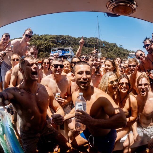 A large group of excited people in swimsuits gathers on a boat, posing cheerfully for the camera. They're under sunny, clear skies, with trees and more boats visible in the background. Everyone appears to be having a fun time.