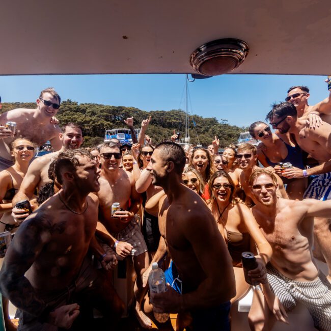 A large group of people in swimsuits are celebrating on a boat. They are smiling, raising drinks, and appear to be enjoying a sunny day on the water. Trees are visible in the background.