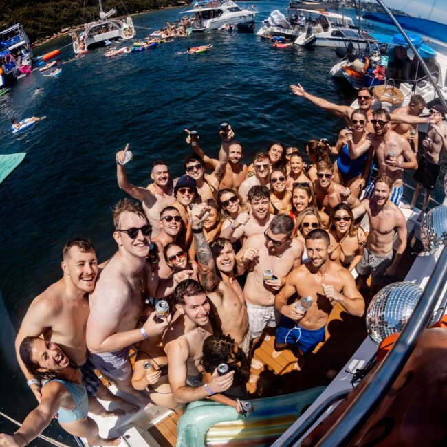 A large group of people in swimwear stand on a yacht, smiling and posing for a photo. The yacht is surrounded by water and several other boats. Some people in the background are swimming and using floating devices. The sky is clear, and the weather is sunny.