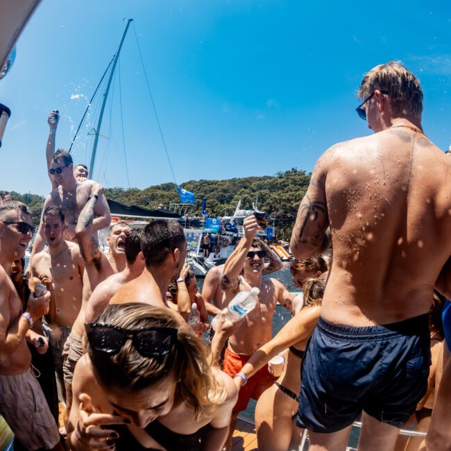 A lively group of people in swimsuits are enjoying a party on a boat under a clear blue sky. Some are dancing, others hold drinks, and one person is pouring water over another's head. Surrounding them are other boats and green, tree-covered hills.