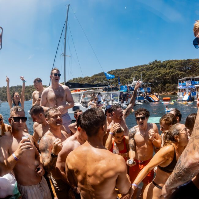 A group of people in swimsuits enjoy a lively party on a boat under a clear blue sky. Some hold drinks, others dance or raise their hands. More boats and a forested shoreline are visible in the background.