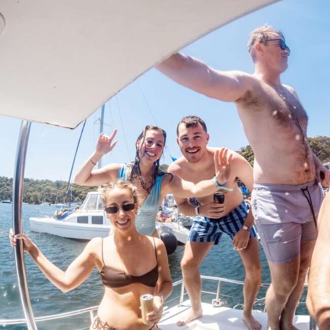 A group of four people enjoying a sunny day on a boat. Two are smiling at the camera, one wearing sunglasses, and another waving. They are in swimwear, with water and other boats visible in the background.