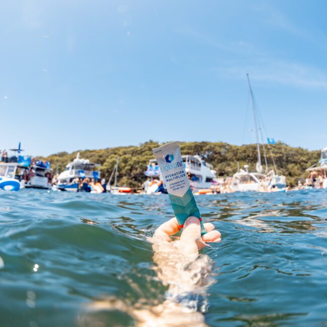 Hand holding a drink in the water, surrounded by boats and people swimming in the background. Sunny day with blue sky.