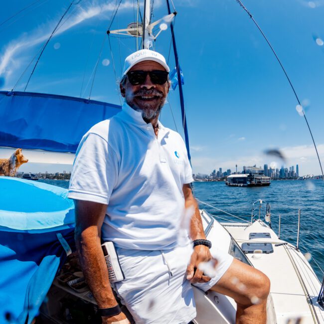 A person wearing a white shirt, shorts, and cap stands on a sailboat, smiling. The ocean and city skyline are visible in the background. It's a sunny day with a blue sky.