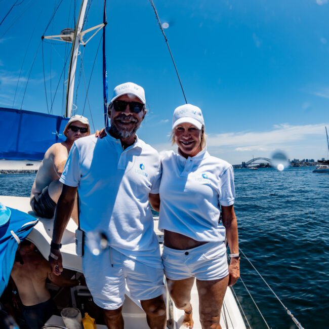 A man and woman in matching white outfits and hats stand on a sailboat under a clear blue sky. The man has sunglasses and a beard. Another person is partially visible behind them. A bridge can be seen in the background over the water.