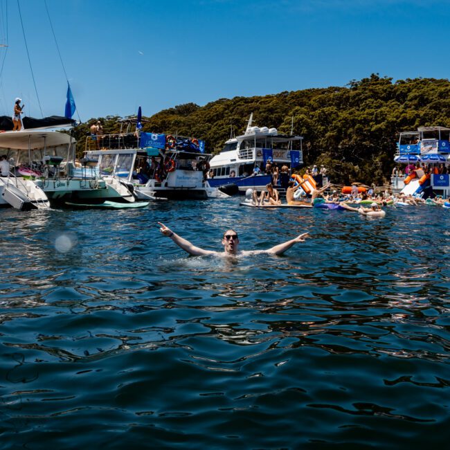 A person is swimming in clear blue water, arms raised. In the background, several boats and yachts are anchored, with people enjoying themselves on board and in the water. The scene is sunny, with lush green trees in the background.