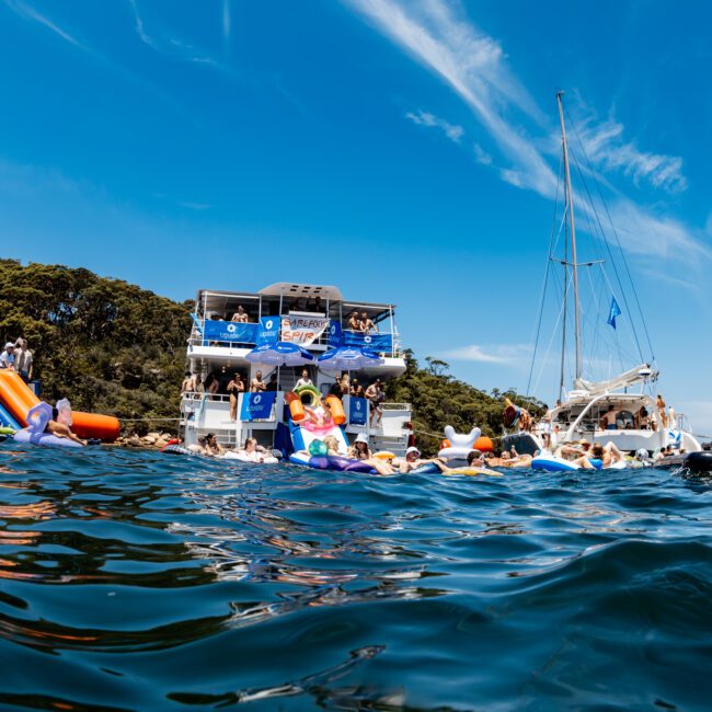 A lively scene of people enjoying a yacht party on the water under a bright blue sky. Several yachts are docked side by side, with party-goers swimming and lounging. Inflatable toys and slides are visible, adding to the festive atmosphere.