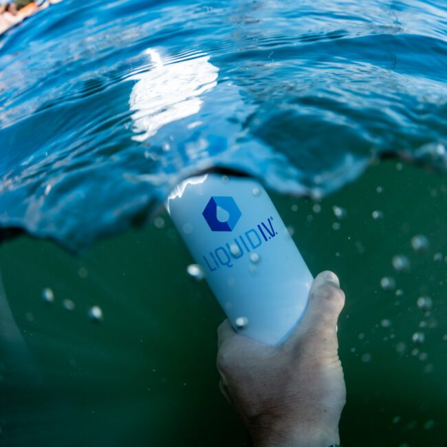 A person holds a white Liquid I.V. bottle partially submerged in ocean water. Boats and a clear sky are visible in the background. The person's wrist is adorned with a silver watch.