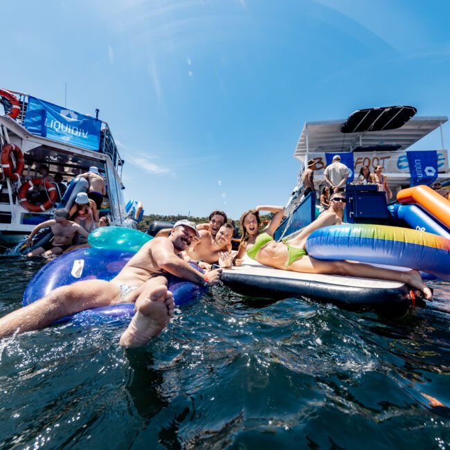 A group of people is enjoying a sunny day on the water, lounging on colorful inflatables between two docked boats. They're smiling, taking a selfie, with the backdrop of a clear sky and lush greenery.