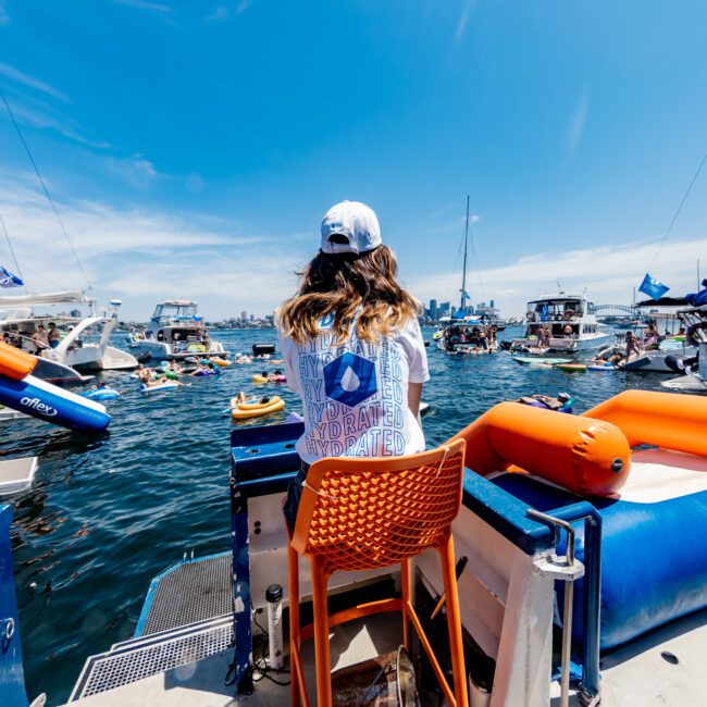 A person sits on an orange chair at the back of a boat, facing a lively scene of numerous boats and people enjoying the water under a clear blue sky. Bright inflatables and city skyline visible in the background.