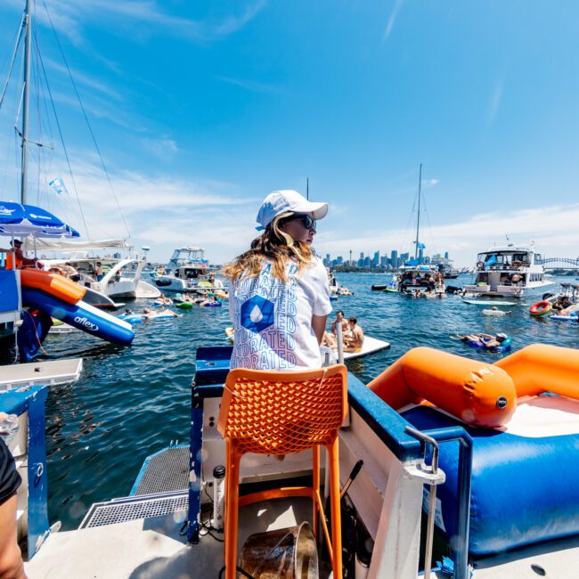 A woman wearing a cap and white T-shirt sits on an orange chair on a boat, facing a group of yachts and people swimming in a sunny harbor. The sky is clear, and city buildings are visible in the distance.