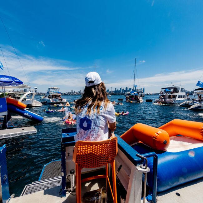 A person in a white cap and life vest sits on an orange chair on a boat, overlooking a lively scene of people enjoying the water. Boats and inflatables are visible under a bright blue sky with scattered clouds.