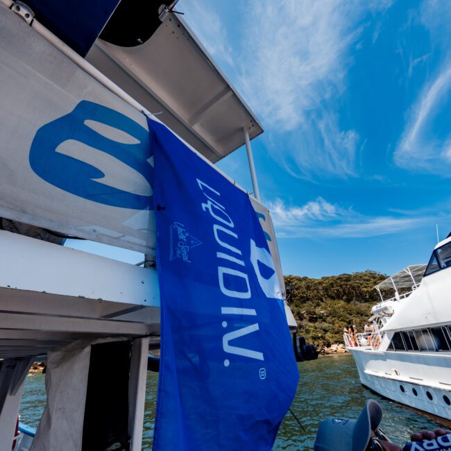 Blue Liquid I.V. flag on a boat with white railings, against a bright blue sky with wispy clouds. Another boat is visible on the right, and there are trees on the distant shore.