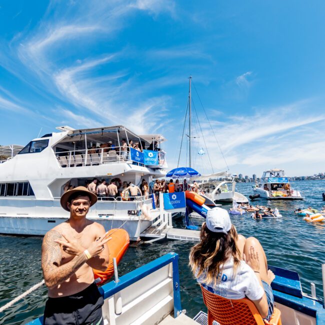 A man in swim trunks and a hat sits on a boat railing holding an orange lifebuoy. A woman in a swimsuit and cap sits nearby. Multiple boats and people are in the water under a sunny blue sky with wispy clouds.