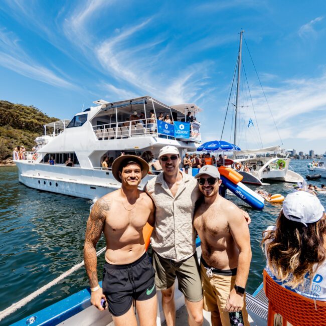 Three people in swimwear stand on a boat dock, smiling. A large yacht filled with people is in the background, along with smaller boats and colorful inflatables on the water, under a clear blue sky. The scene is lively and festive.