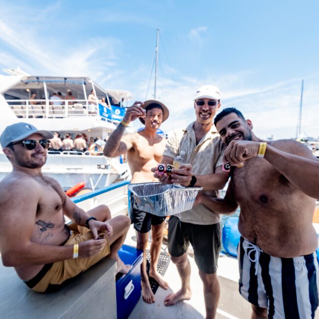 Four men on a boat, smiling and holding drinks. They are shirtless except one in a light shirt. Behind them is a large boat with people onboard. The water and city skyline are visible in the background. It's a sunny day.