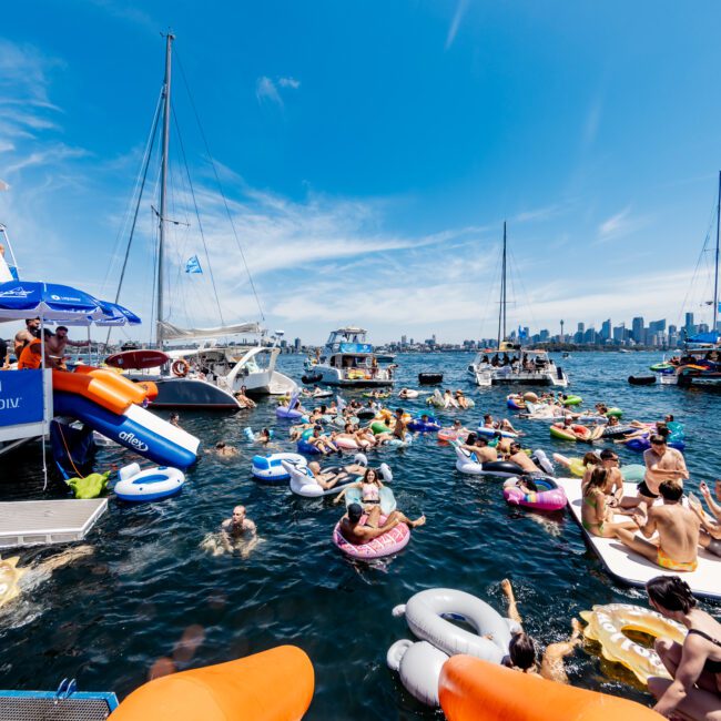 A vibrant scene on the water with numerous people enjoying a sunny day on inflatable rafts. Several boats are anchored nearby, and the city skyline is visible in the background. The atmosphere is lively and festive.