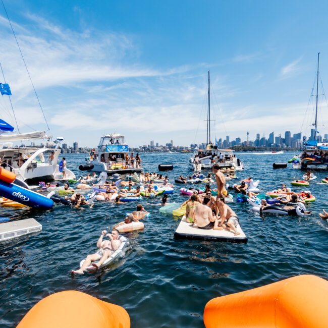 A lively scene on the water with people enjoying a sunny day on floaties near boats. The city skyline is visible in the background. Bright orange inflatable slides are in the foreground.