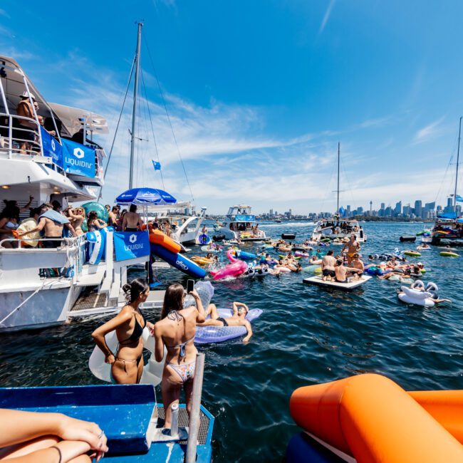 People relax on colorful inflatables in the water near boats, under a clear blue sky. The city skyline is visible in the distance. Some individuals are on a yacht with a logo that says "Liquid.