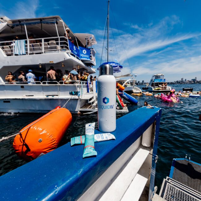 A lively scene on the water with boats and people swimming and lounging on floaties. Foreground features a white water bottle and sunscreen on a boat railing. Bright sunny day with a few clouds in the sky.