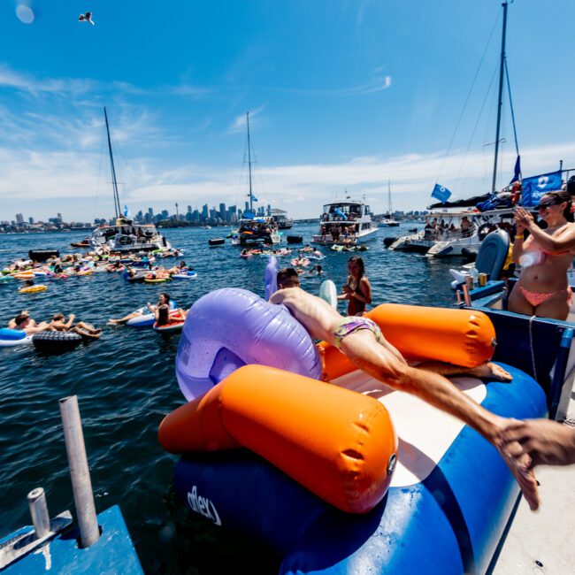 People are enjoying a sunny day on the water. A person dives from a floating platform into the sea, surrounded by boats, inflatables, and others in swimwear. The city skyline is visible in the background under a clear blue sky.