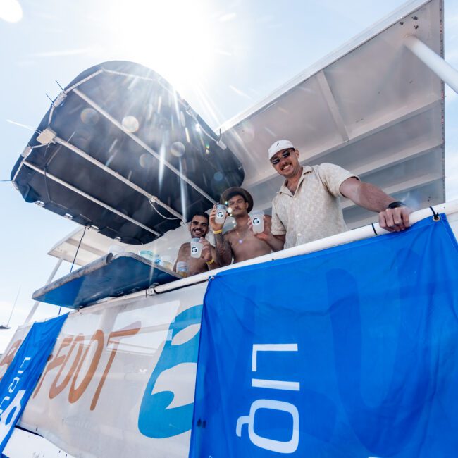 Four people smiling and holding cans on the upper deck of a boat, with blue banners hanging below. Sunlight shines from the top left, creating a bright atmosphere.